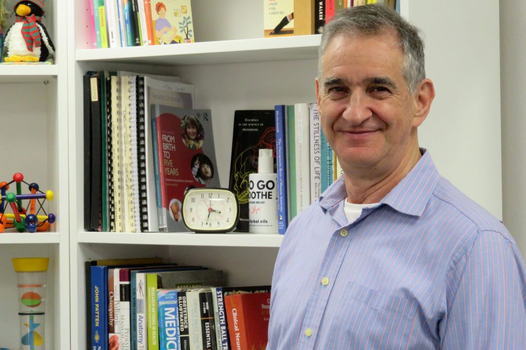 East Devon Osteo Joyaa Antares standing in front of bookcase in his office