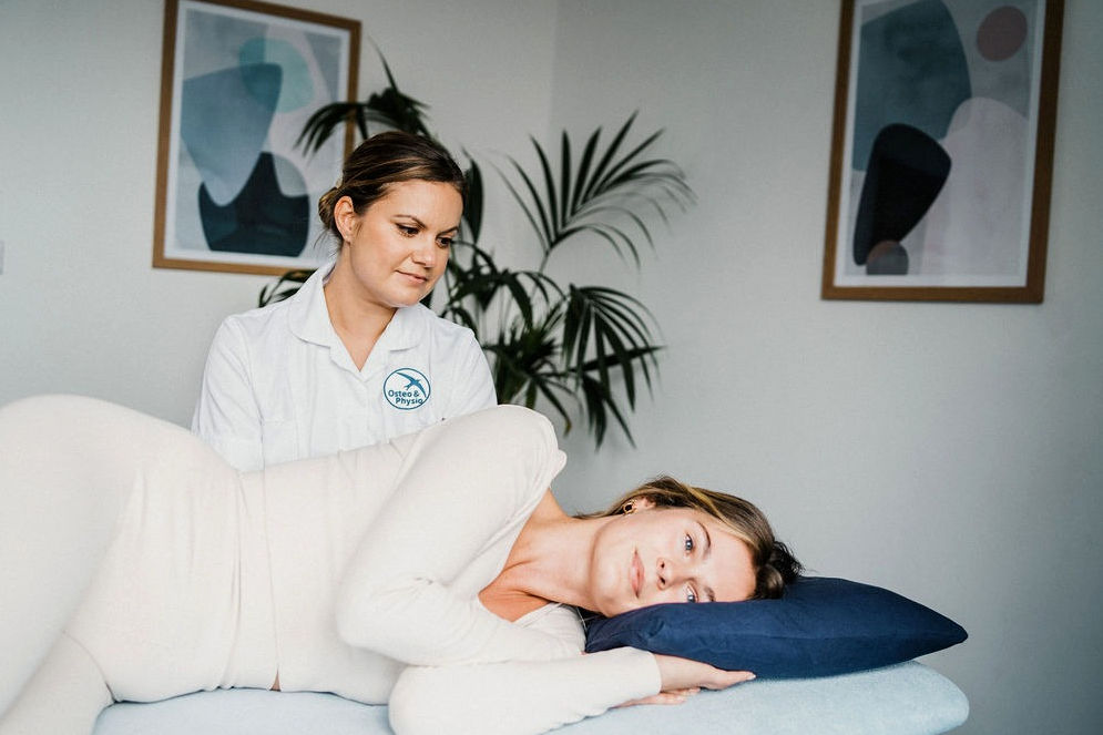A woman lies on her side on an exam table with her head on a pillow while a Osteo & Physio staff member observes her